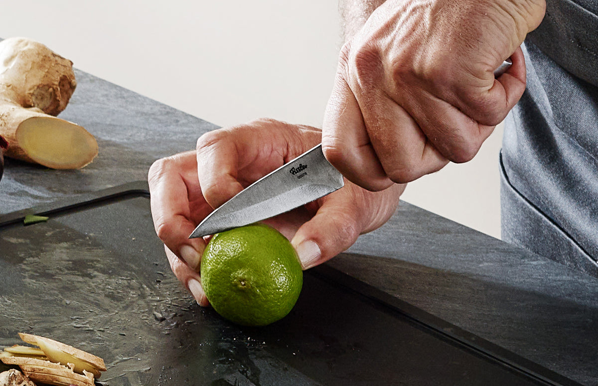 Eine Person schneidet eine Limette mit dem Fissler Essential® Schälmesser auf einer glatten Steinoberfläche; im Hintergrund liegt eine frische Ingwerwurzel.