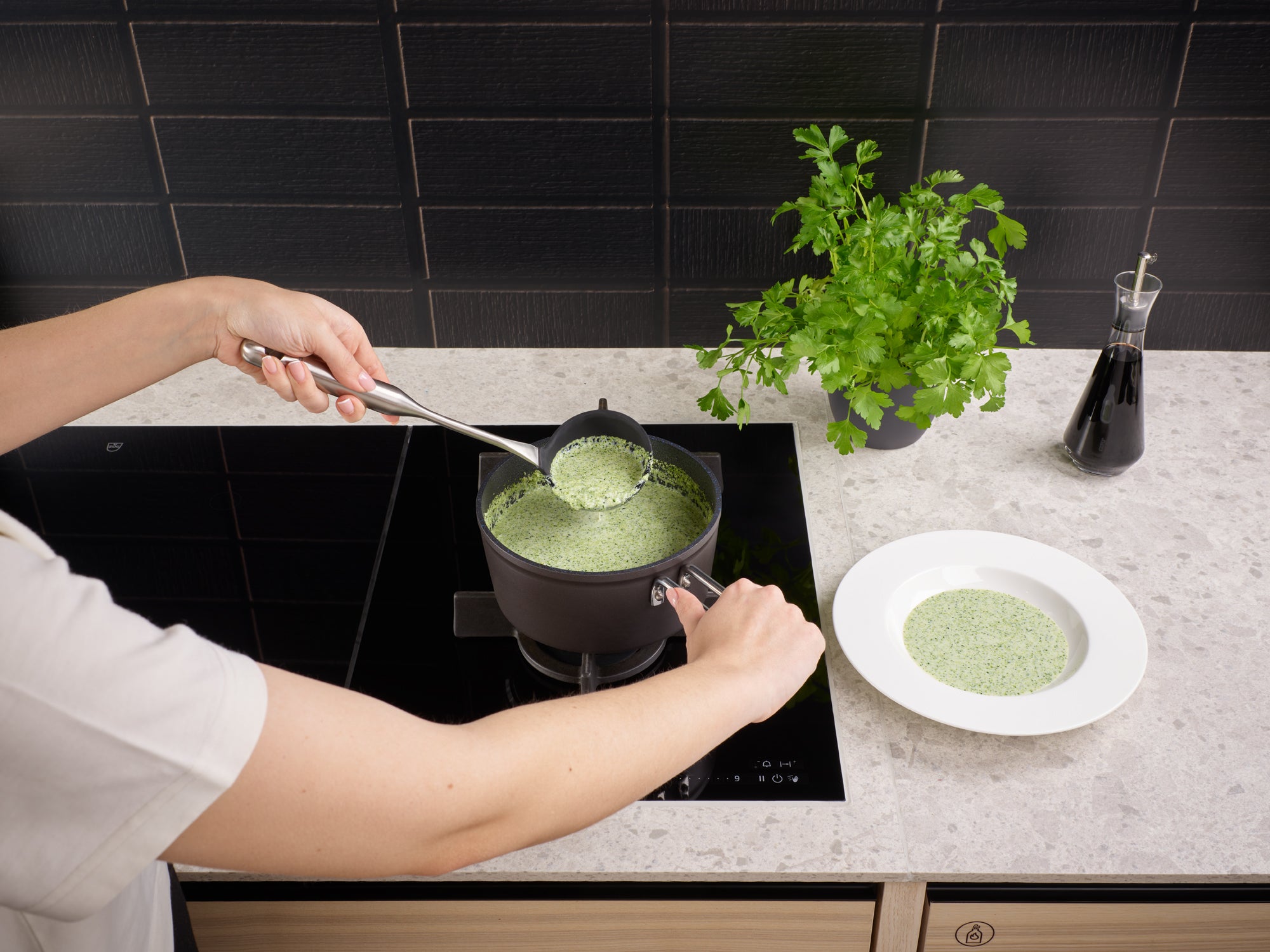 Eine Person serviert eine Kräutersuppe aus einer Fissler Adamant® Stielkasserolle auf einem Induktionskochfeld und unterstreicht damit deutsche Präzision und Design.