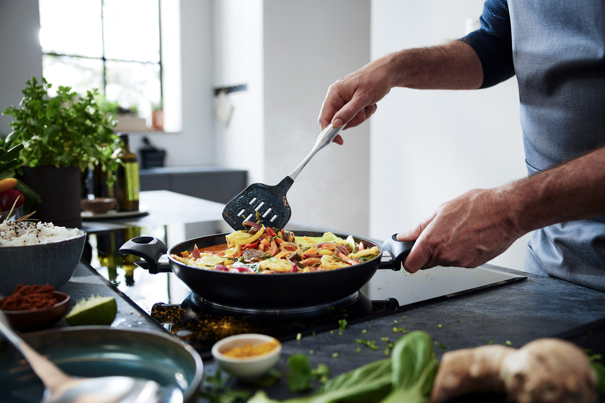 Kochen von lebhaftem Gemüse in einer schwarzen Fissler Essential® Pfanne mit Bratwender mit Silikon, frische Kräuter auf der Theke.