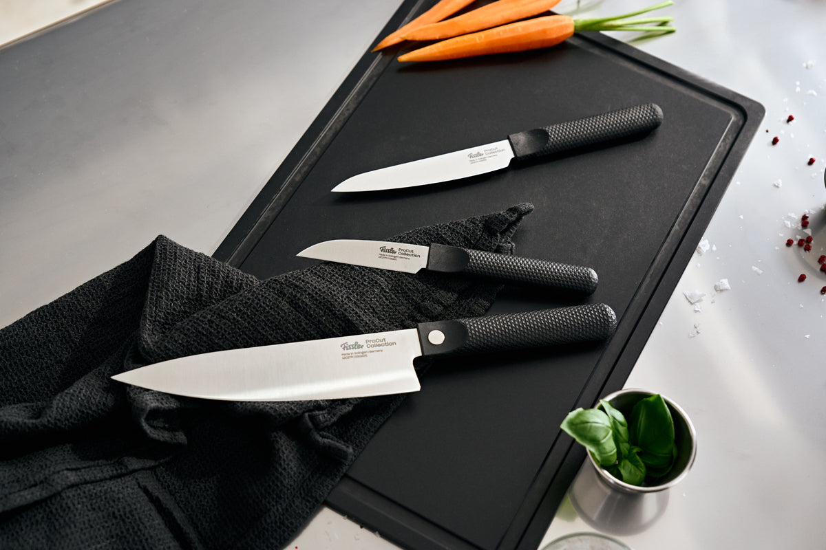 Fissler ProCut Messerset mit schwarzen Griffen auf Schneidebrett, umgeben von Karotten, Basilikum und Gewürzen.