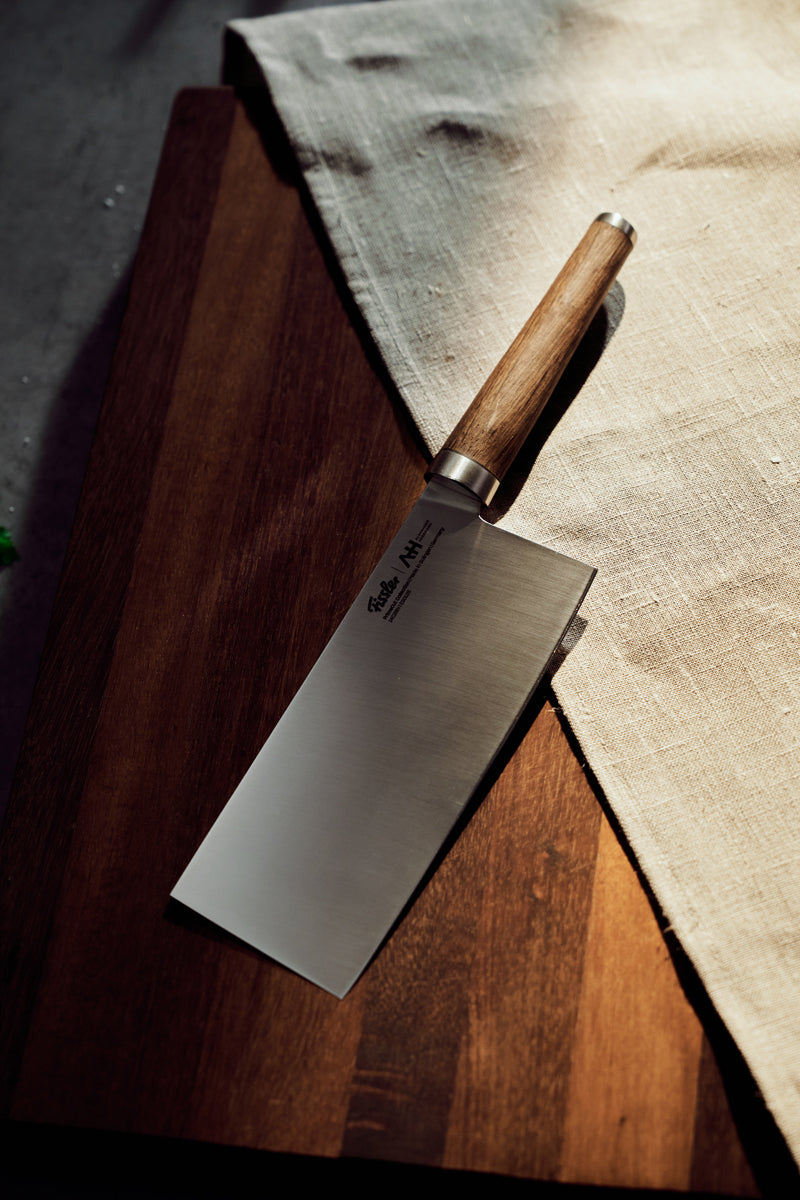 Fissler PrimeCut Chinesisches Kochmesser 18cm mit Holzgriff auf einem dunklen Brett und beigem Tuch, in warmem Licht. Hergestellt in Deutschland.