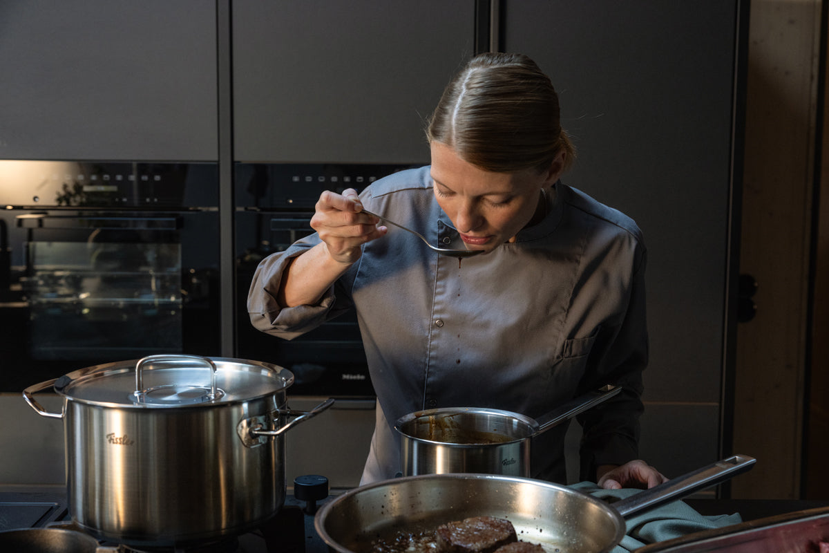 A chef in a gray uniform tastes food on a modern kitchen stove next to Fissler Original-Profi Collection® stainless steel pots.