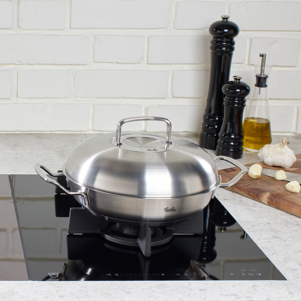 Fissler Original-Profi Collection® Servierpfanne mit Novogrill®-Deckel auf Gasherd, umgeben von Salz, Pfeffer und Olivenöl.