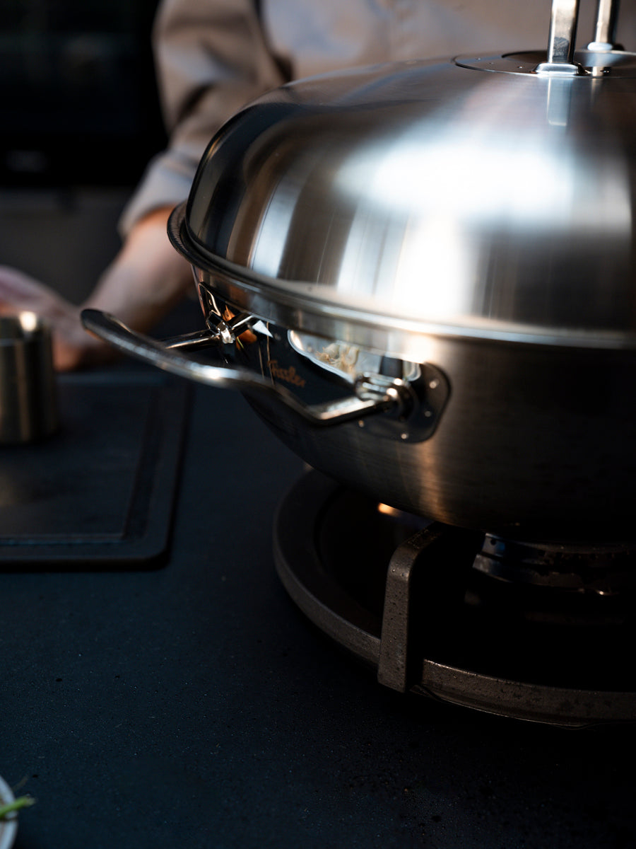 Fissler Original-Profi Collection® Wok 32cm mit gewölbtem Deckel, präzisionsgefertigt in Deutschland, auf glatter Oberfläche; Person dahinter verschwommen.