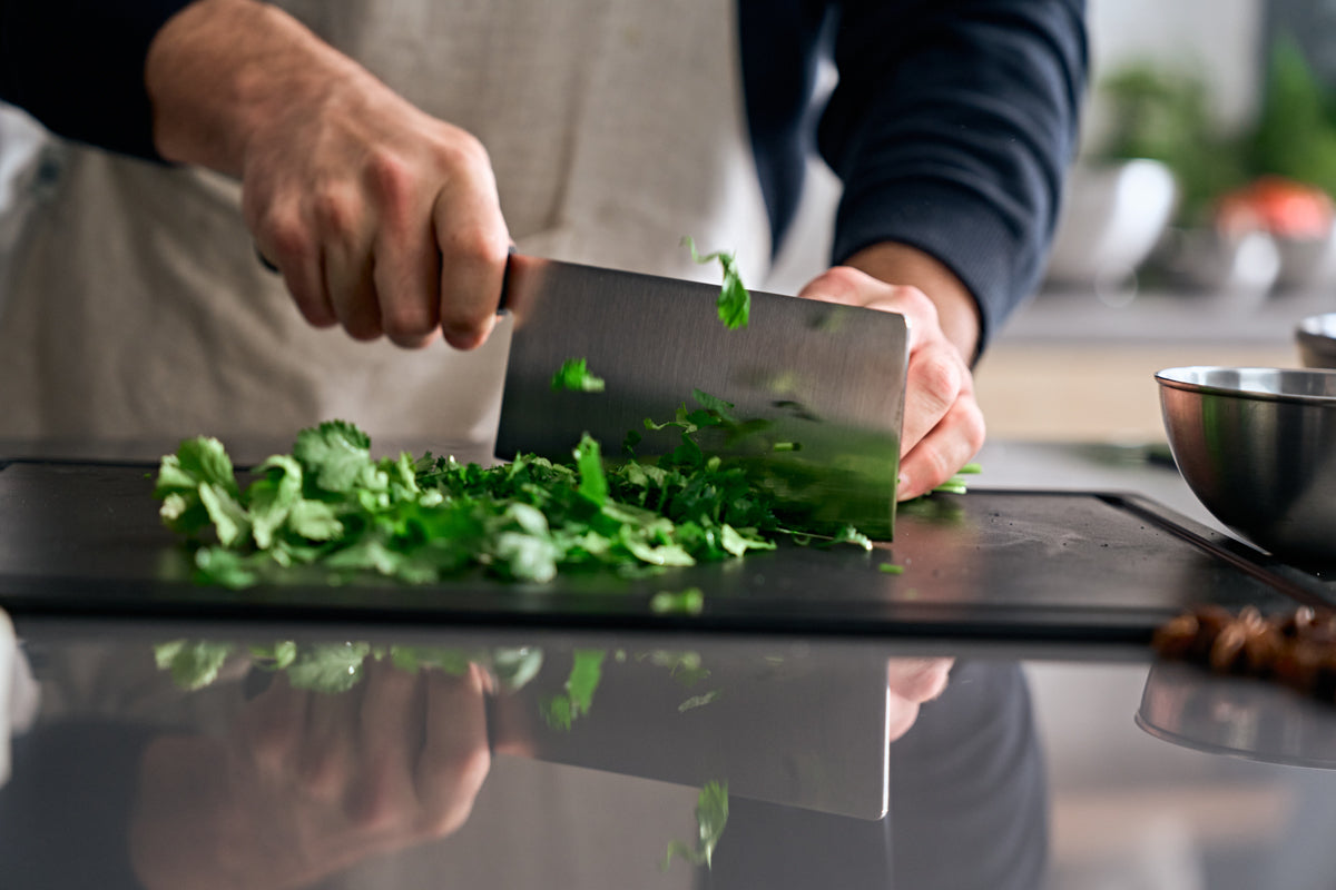Eine Person schneidet Kräuter mit dem Fissler ProCut Chinese Chef's Knife auf einem schwarzen Brett, das deutsche Präzision und modernes Design zeigt.