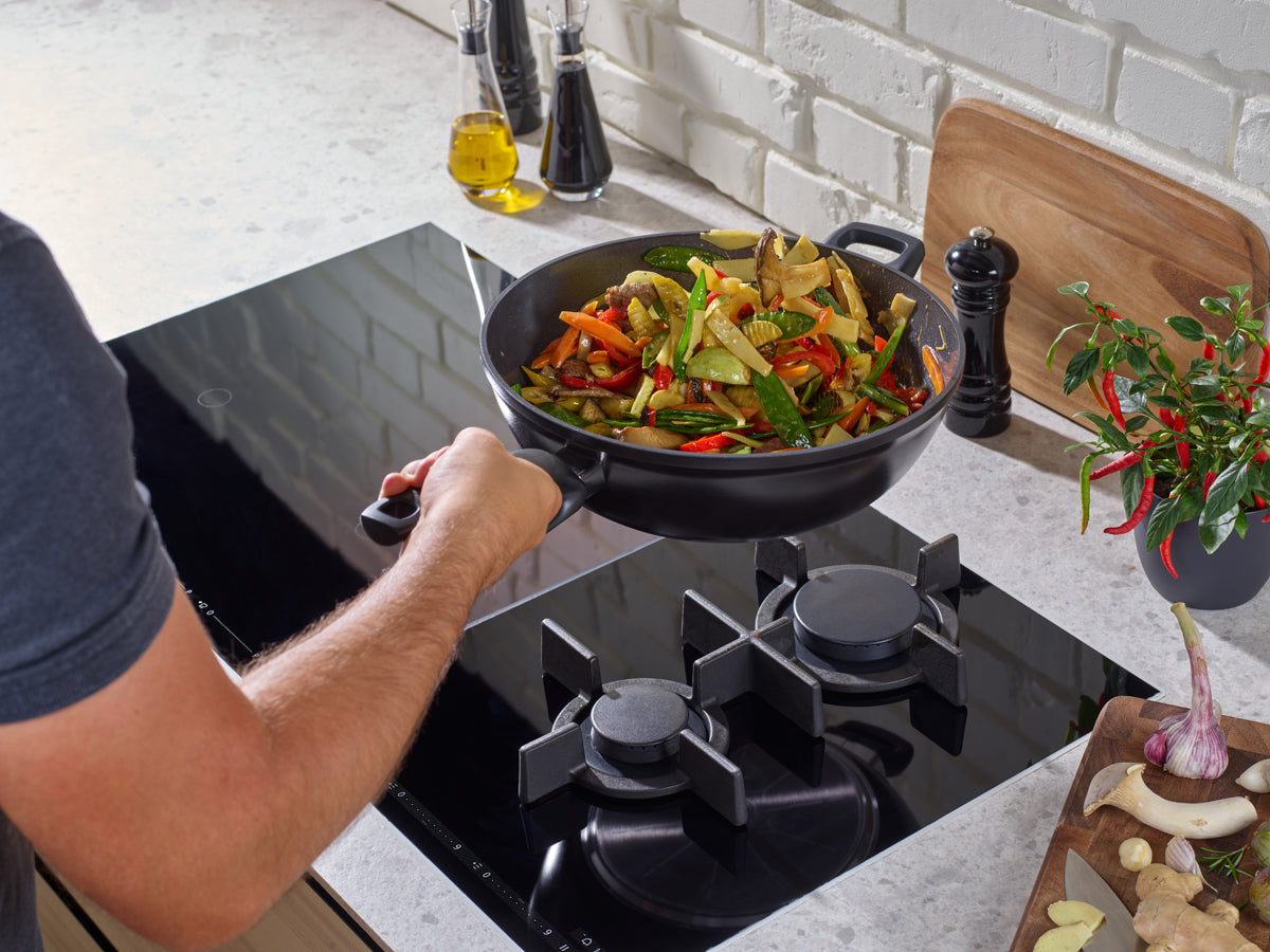 In einem Fissler Ceratal® Wok Orbitschwarz wird ein Gemüsepfannengericht zubereitet; Gewürze und frische Zutaten liegen auf einer schlichten, modernen Theke.
