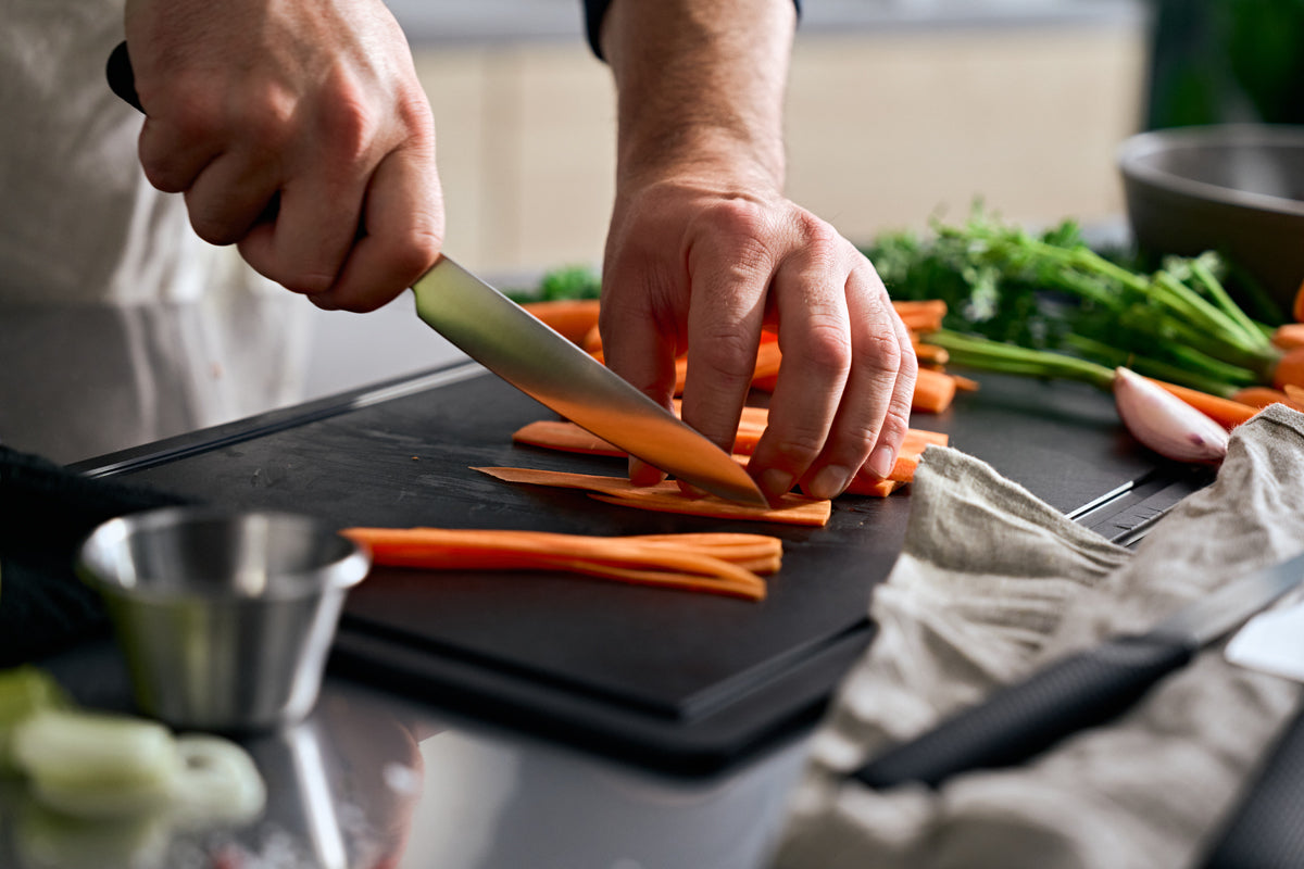 Eine Person schneidet Karotten auf einem schwarzen Brett mit dem Fissler ProCut Universalmesser 14cm; andere Fissler-Messer und Kräuter in der Nähe.
