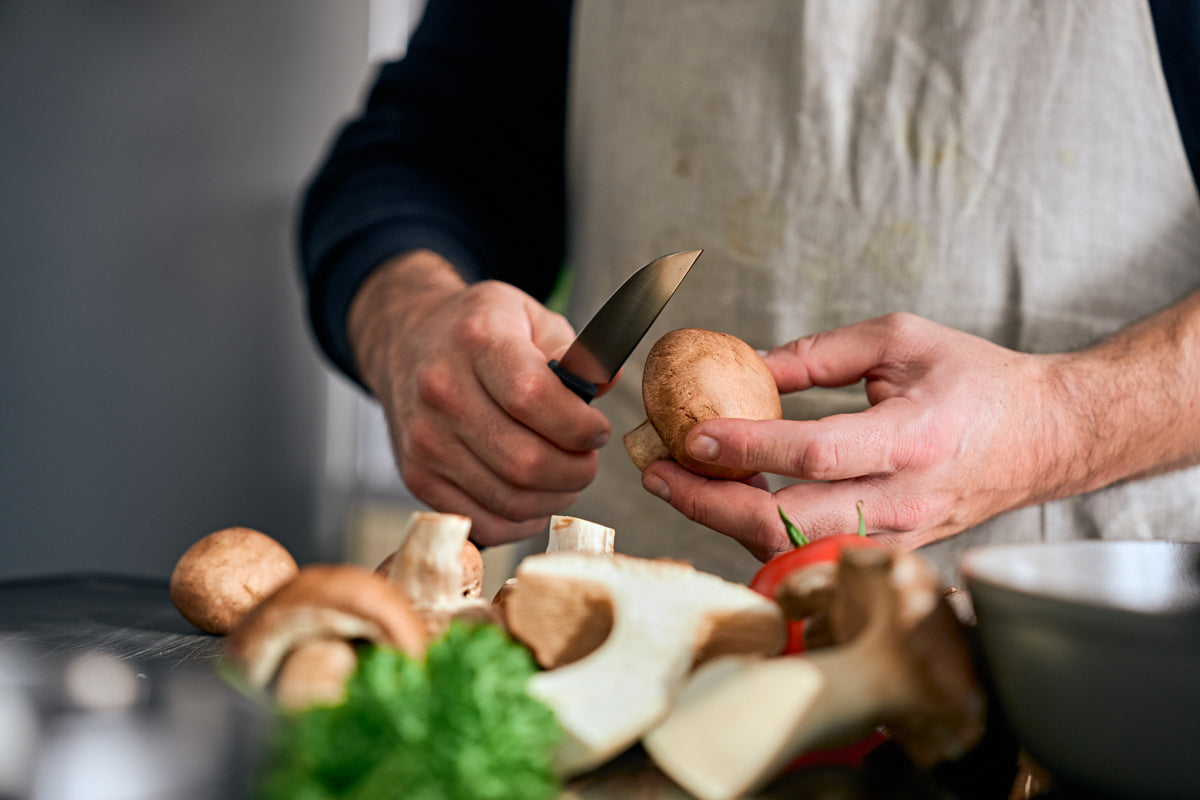 Eine Person in einer Schürze schneidet einen Pilz mit dem Fissler ProCut Gemüsemesser 9cm, mit frischen Zutaten auf einer modernen Theke.