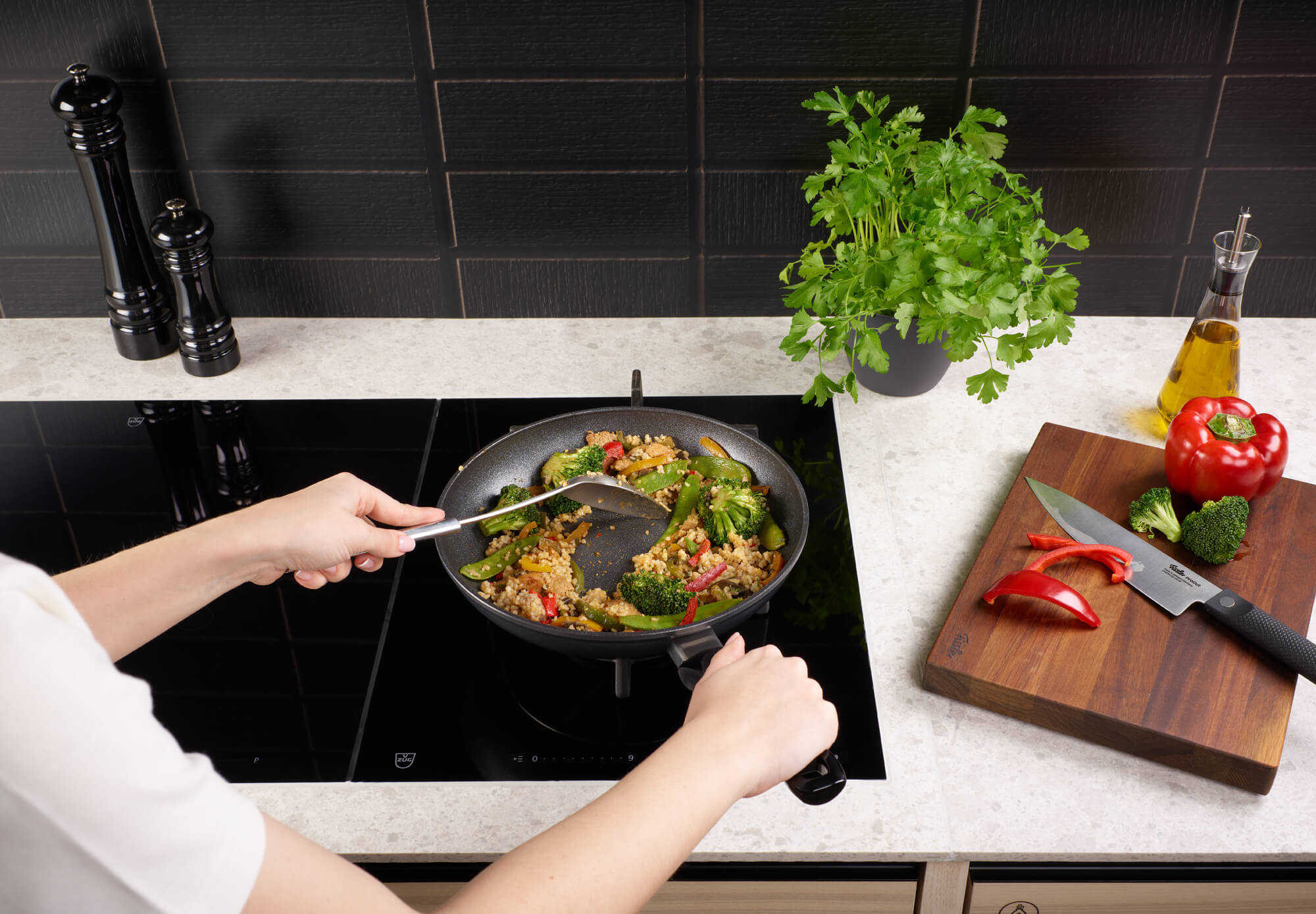 Eine Person kocht mit der Fissler Adamant® Classic Pfanne auf dem Herd, umgeben von frischen Kräutern, Gemüse und Küchengeräten.