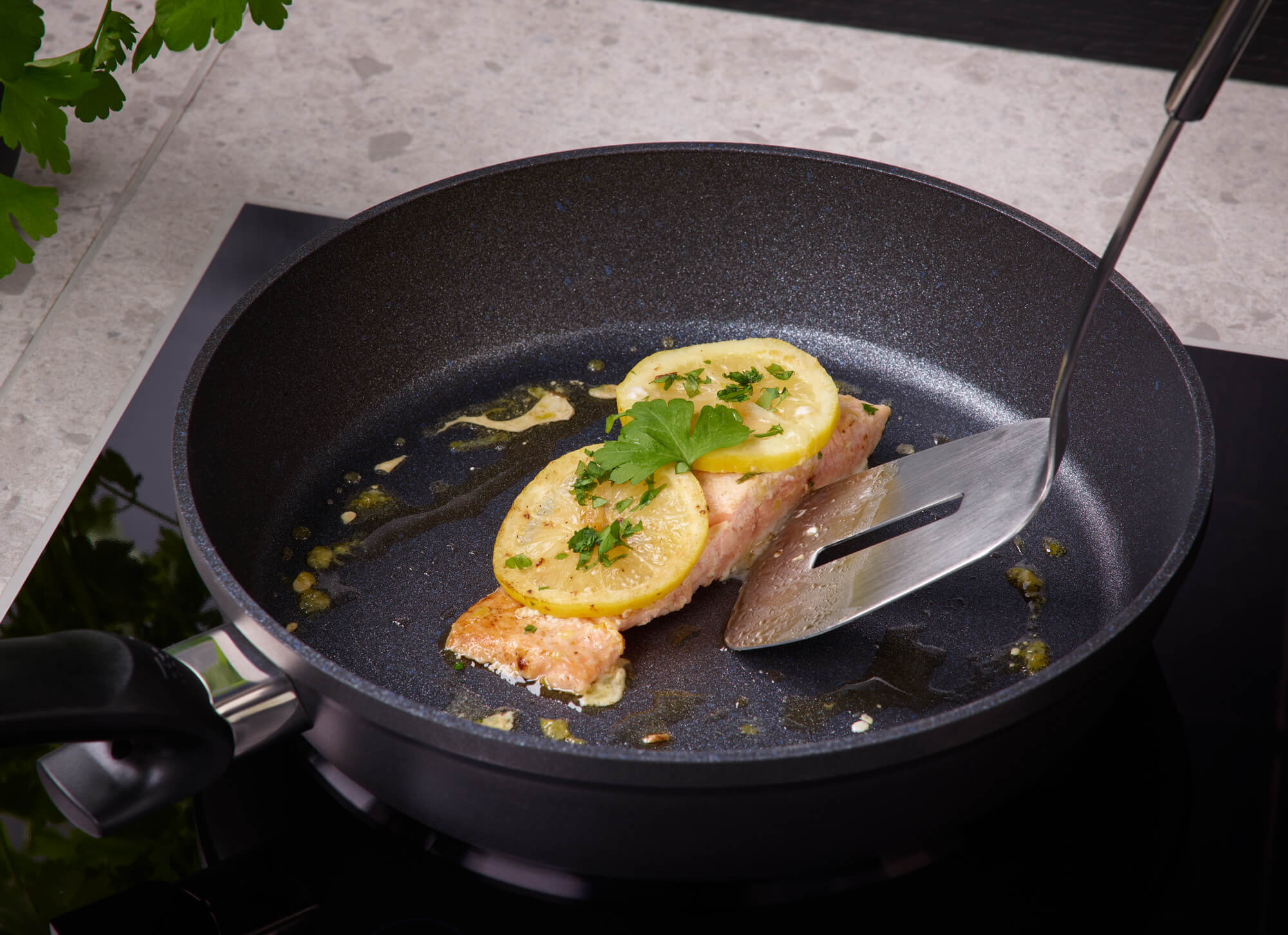 Salmon fillet with lemon and herbs in a Fissler Adamant® Comfort Frying Pan, lifted with a spatula; fresh parsley in the background.