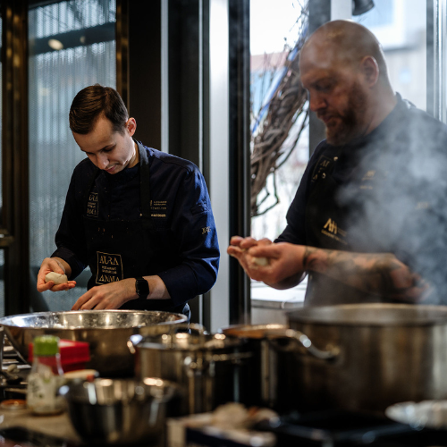 Two chefs in navy uniforms prepare food in a kitchen equipped with premium Fissler stainless steel cookware. One chef shapes dough beside an array of expertly crafted pots and pans, while the other cooks near steaming Fissler pressure cookers. Precision-engineered kitchen tools and timelessly designed accessories are visible on the counter, showcasing German innovation and durability.