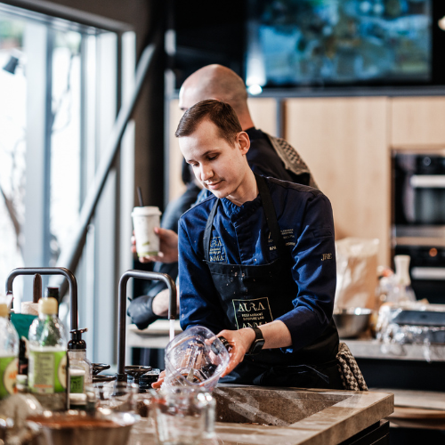 A chef in a blue uniform and apron cleans Fissler stainless steel pots and pans at a modern kitchen counter, surrounded by precision-engineered utensils and premium ingredients. In the background, another person stands holding a drink, highlighting the welcoming atmosphere that Fissler’s timeless cookware brings to every home kitchen.
