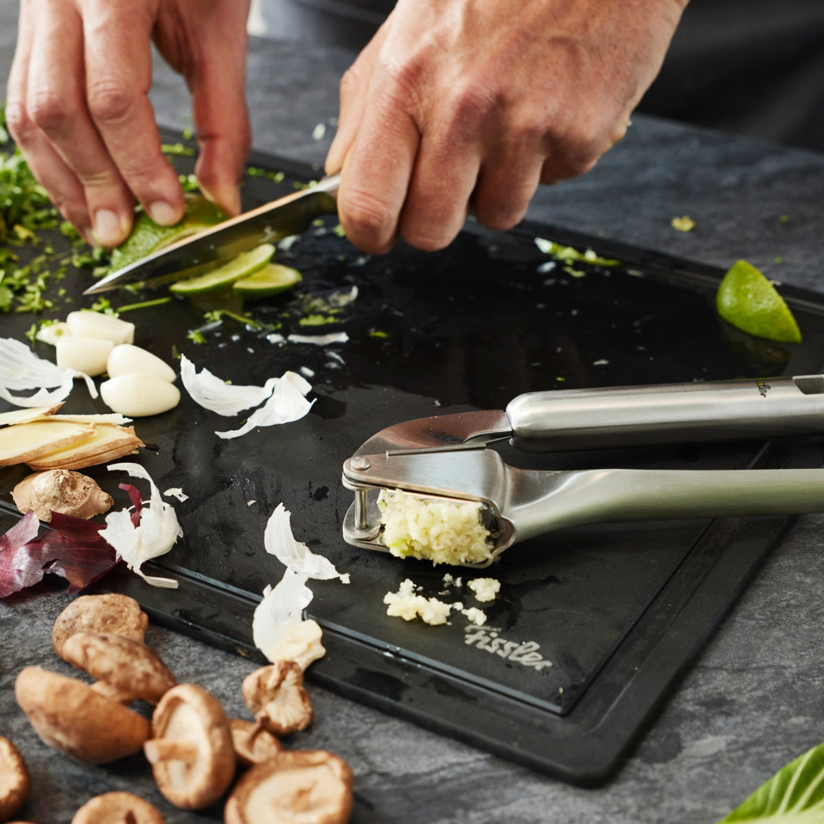 Frische Kräuter und Limetten auf einem schlichten schwarzen Brett mit Knoblauch, Pilzen, Ingwer und der Fissler Essential® Knoblauchpresse.