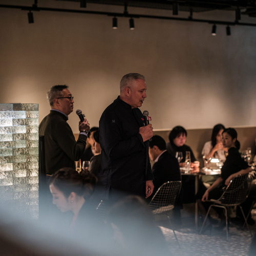 Two men stand speaking into microphones at an event, addressing an audience seated at round tables in a contemporary, softly lit restaurant space. Fissler’s renowned stainless steel cookware and pressure cookers are showcased on elegantly set tables, reflecting the brand’s commitment to precision engineering and timeless design.