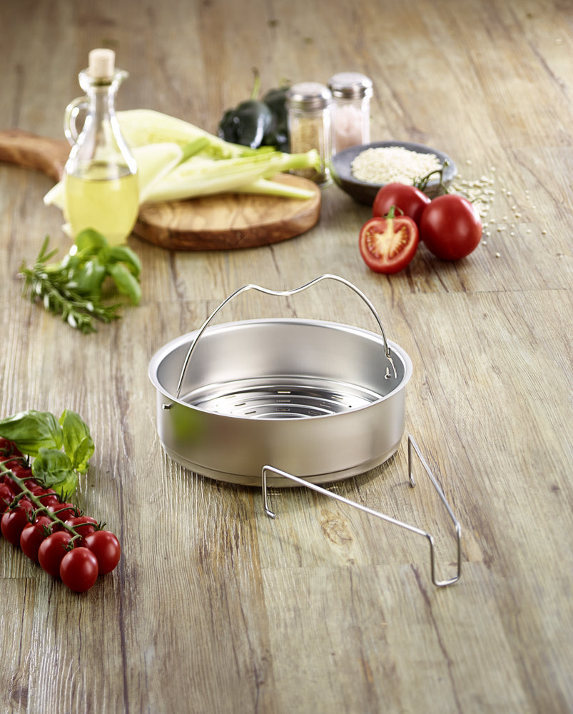 Fissler Pressure Cooker Accessories insert, perforated with handles and tripod, surrounded by fresh vegetables - modern, functional design.