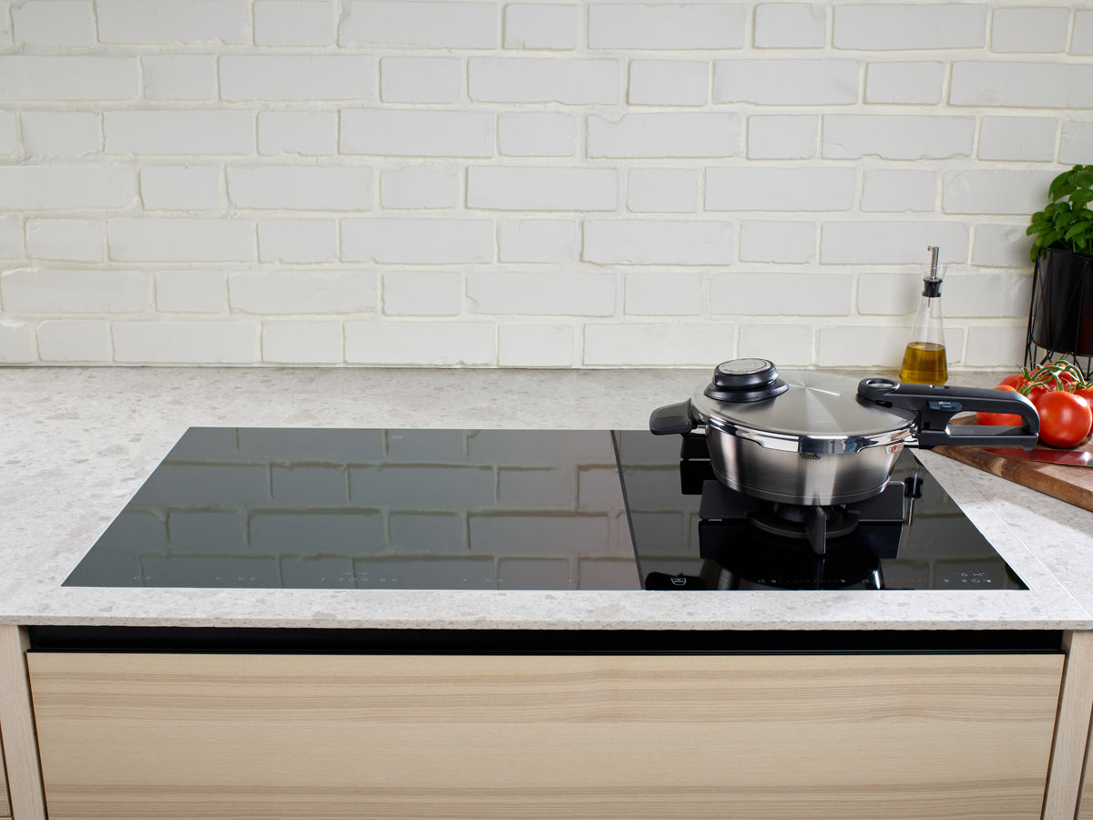 In einer modernen Küche mit einem eleganten Induktionskochfeld aus schwarzem Glas steht die Fissler Vitavit® Premium Schnellbratpfanne aus hochwertigem Edelstahl 18/10 mit ihrem passenden Deckel elegant auf einer Kochstelle. Charakteristische Akzente wie Speiseöl, reife Tomaten und frisches Basilikum liegen auf der Arbeitsplatte vor einer klassischen weißen Ziegel-Fliesenwand – eine einladende Szene, die Fisslers deutsche Präzision und zeitloses Design unterstreicht.