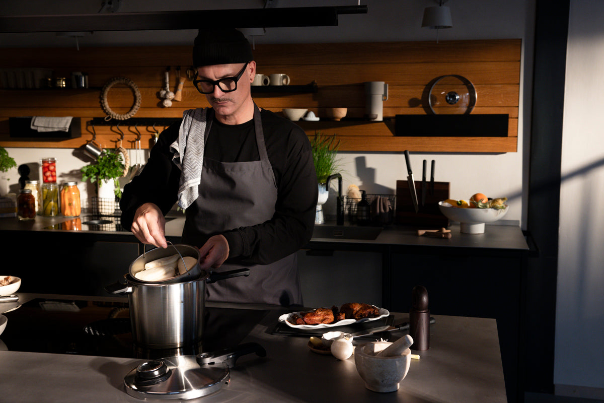 In einer modernen, sonnendurchfluteten Küche rührt eine Köchin in einem Fissler Vitavit® Premium Schnellkochtopfset, Kräuter und Utensilien liegen daneben.
