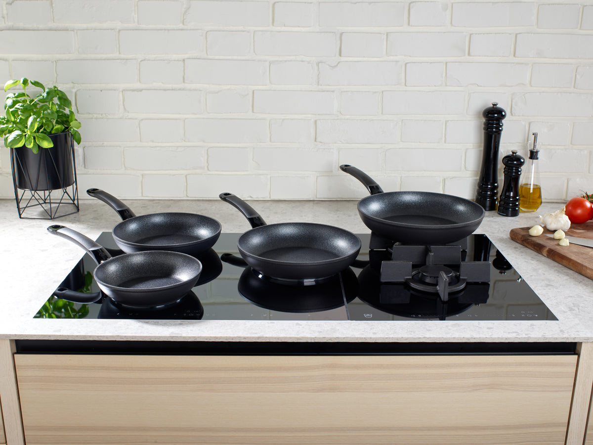 Five Fissler Adamant® Classic pans on a modern stove, showcasing the quality of German manufacturing with herbs and fresh ingredients.