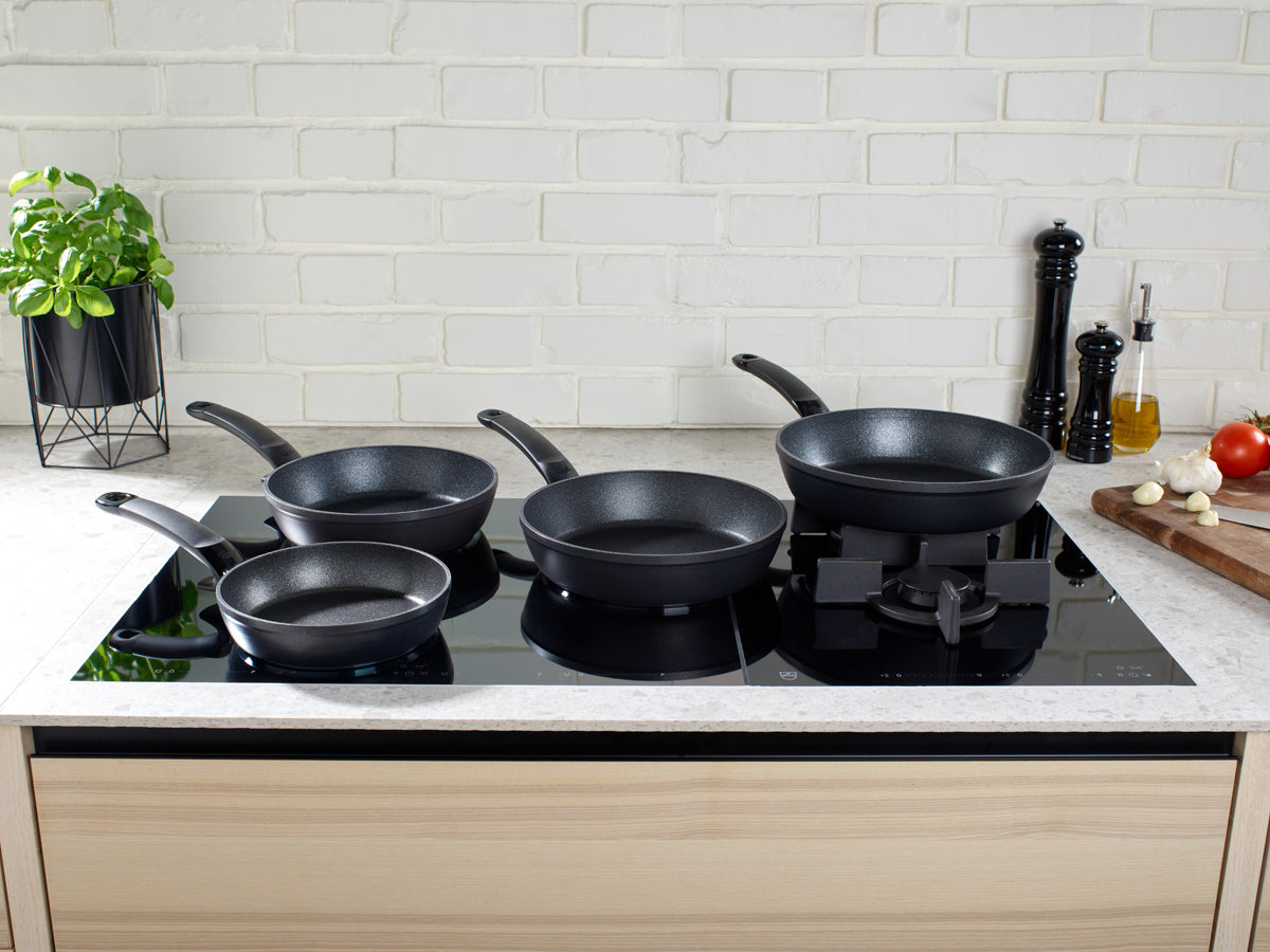 Four Fissler Adamant® Comfort non-stick pans on a modern stovetop, with fresh herbs and vegetables. Precision engineering in Germany.