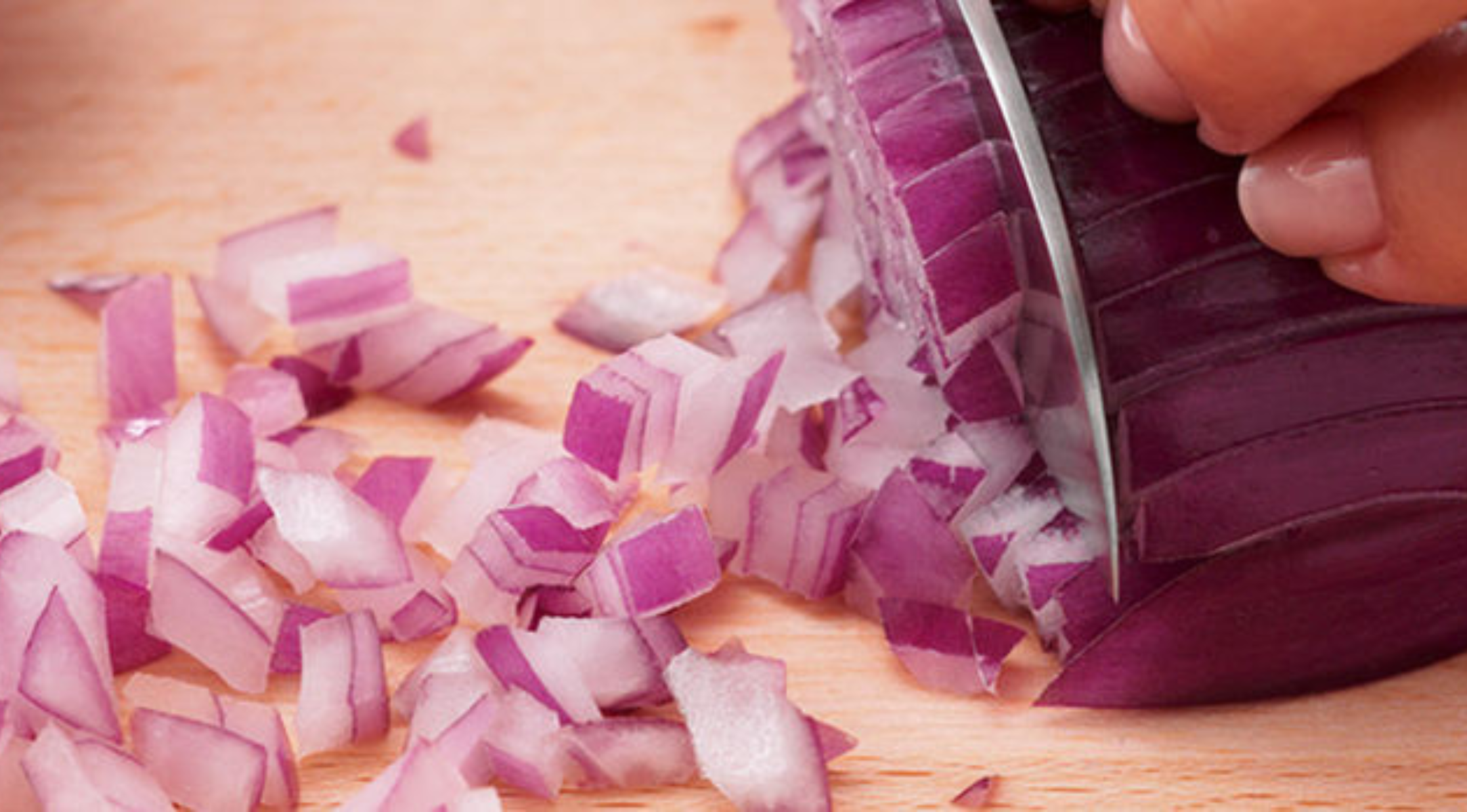 Nahaufnahme von Händen, die eine rote Zwiebel auf einem hölzernen Schneidebrett präzise zerkleinern, ein Beispiel für die deutsche Fissler-Küchenkunst.