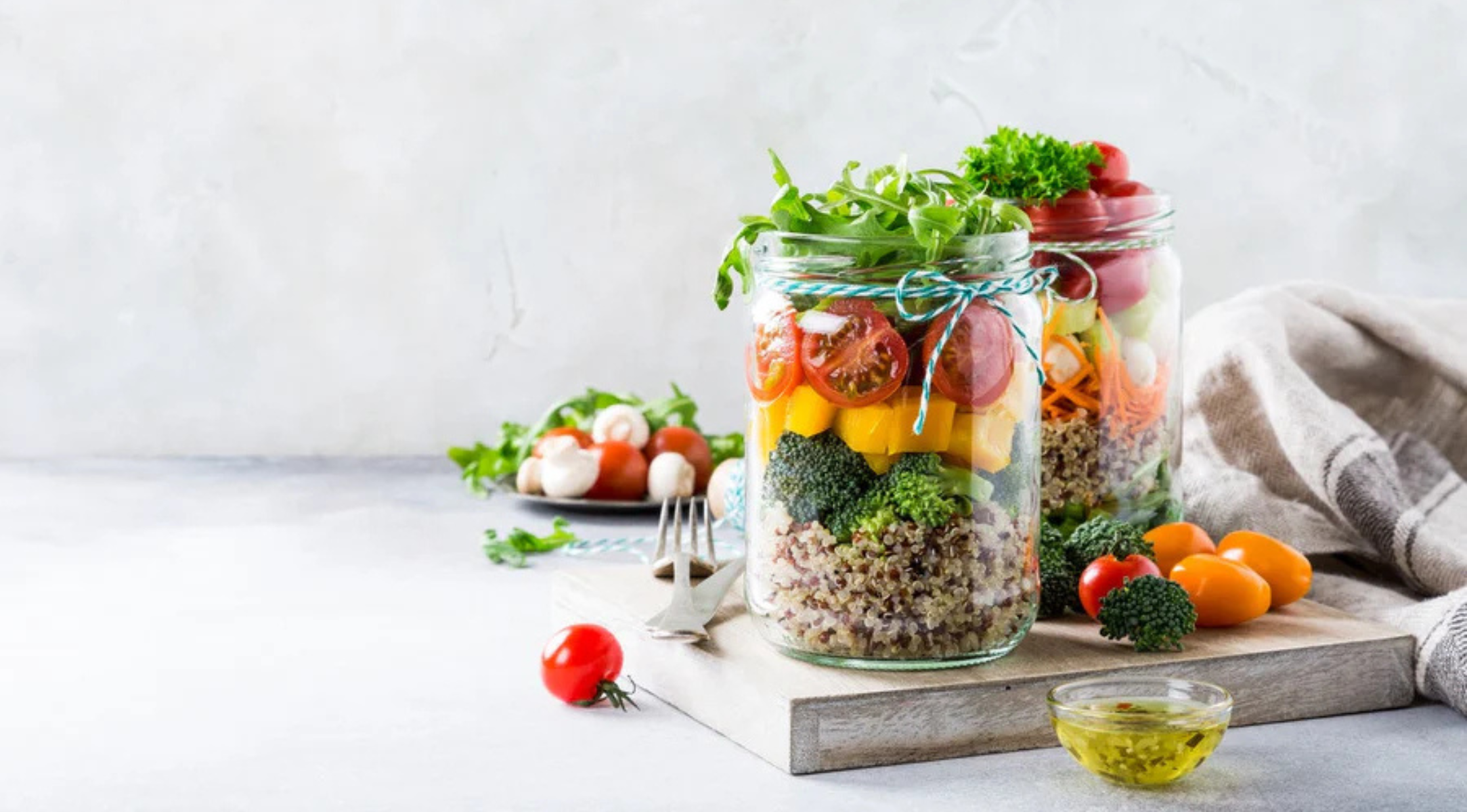 Zwei Gläser mit bunten Salatschichten, frischem Gemüse und Dressing stehen auf einem Holzbrett neben dem Fissler-Kochgeschirr.