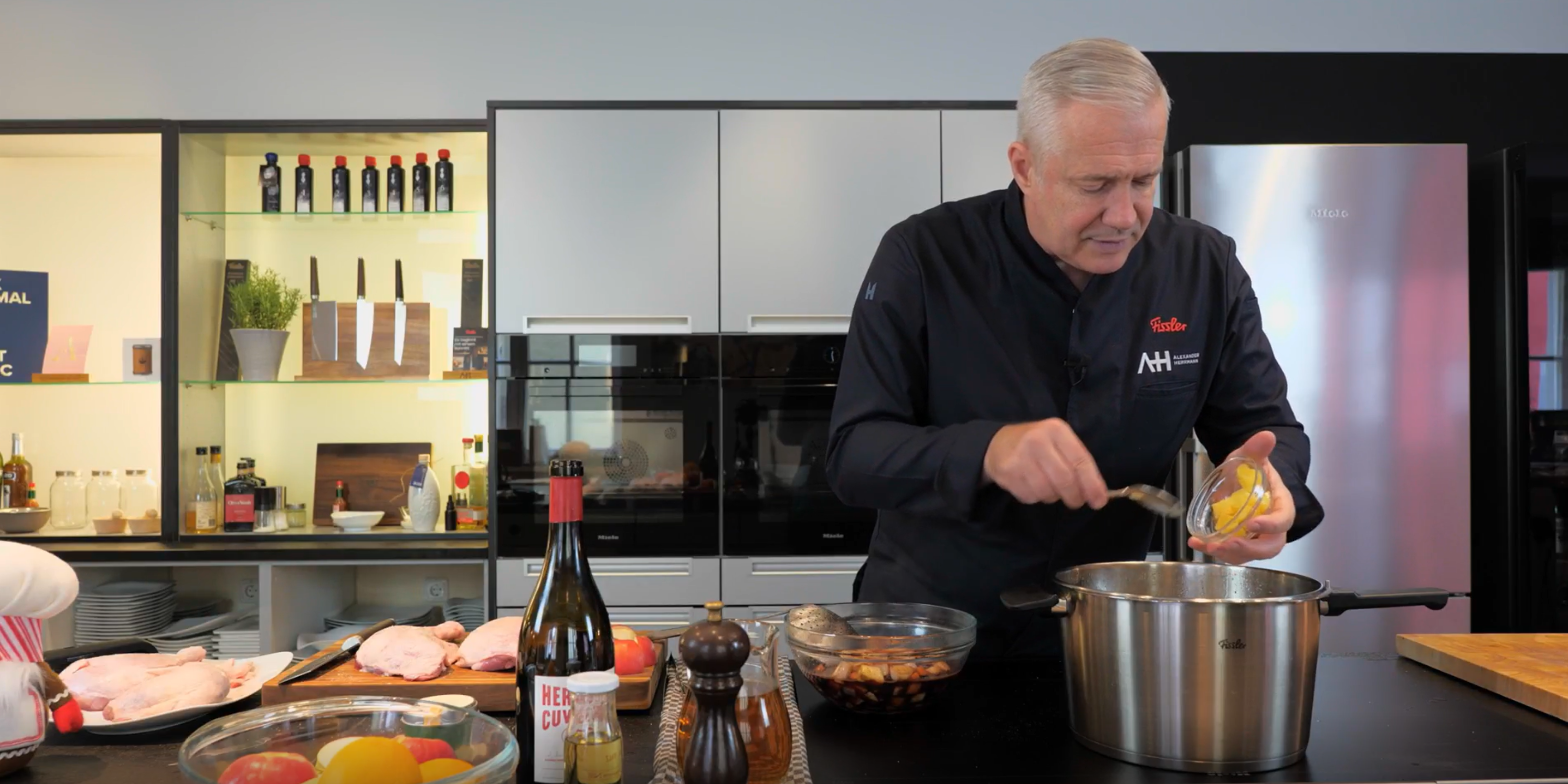 Ein Chefkoch verwendet hochwertiges Fissler-Kochgeschirr in einer eleganten Küche und gibt die Zutaten fachmännisch in einen Edelstahltopf inmitten hochwertiger Utensilien.