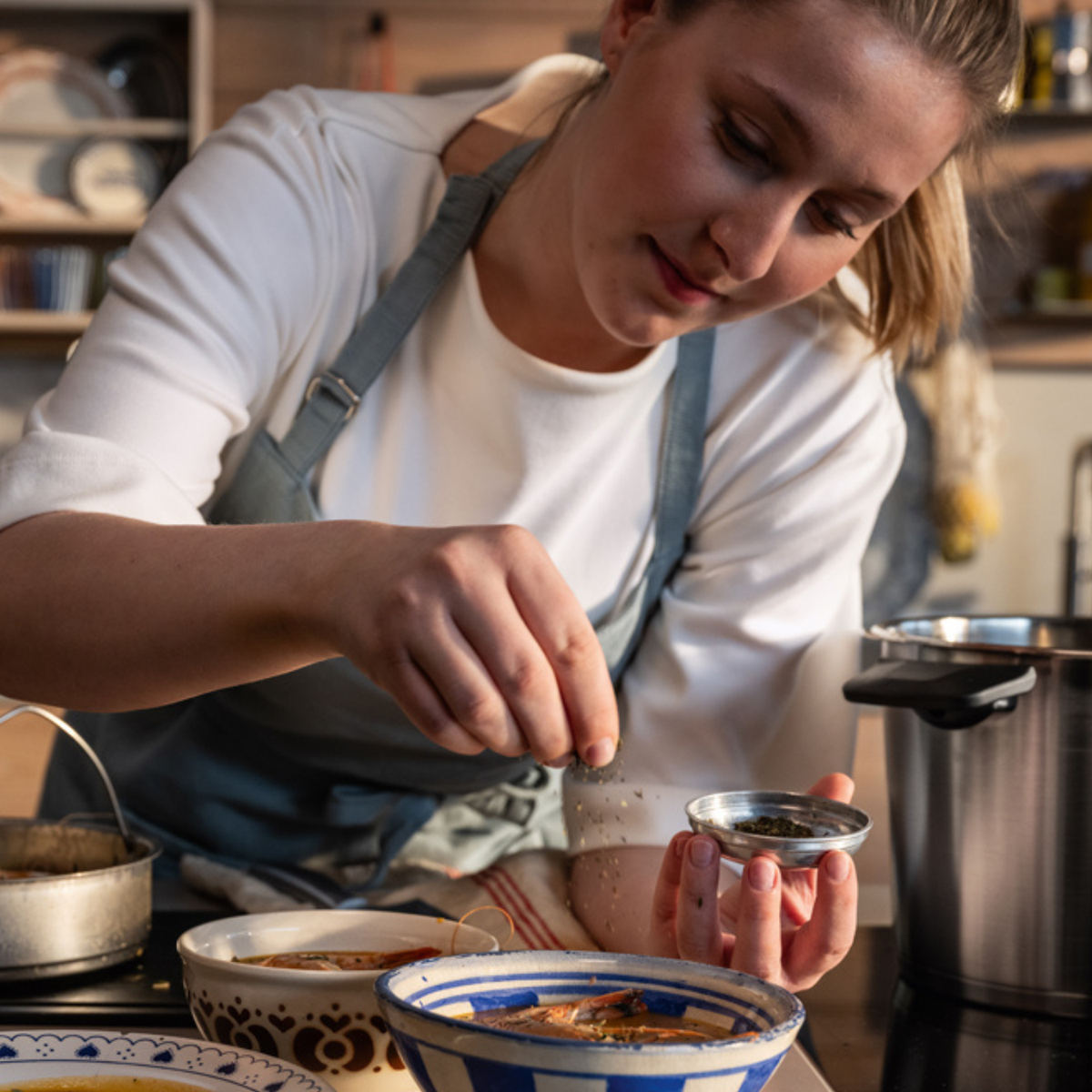 A person in an apron sprinkles seasoning over a bowl of food in a kitchen, surrounded by premium Fissler stainless steel pots and cookware. The scene reflects Fissler’s tradition of precision engineering, timeless design, and innovation “Made in Germany.”.