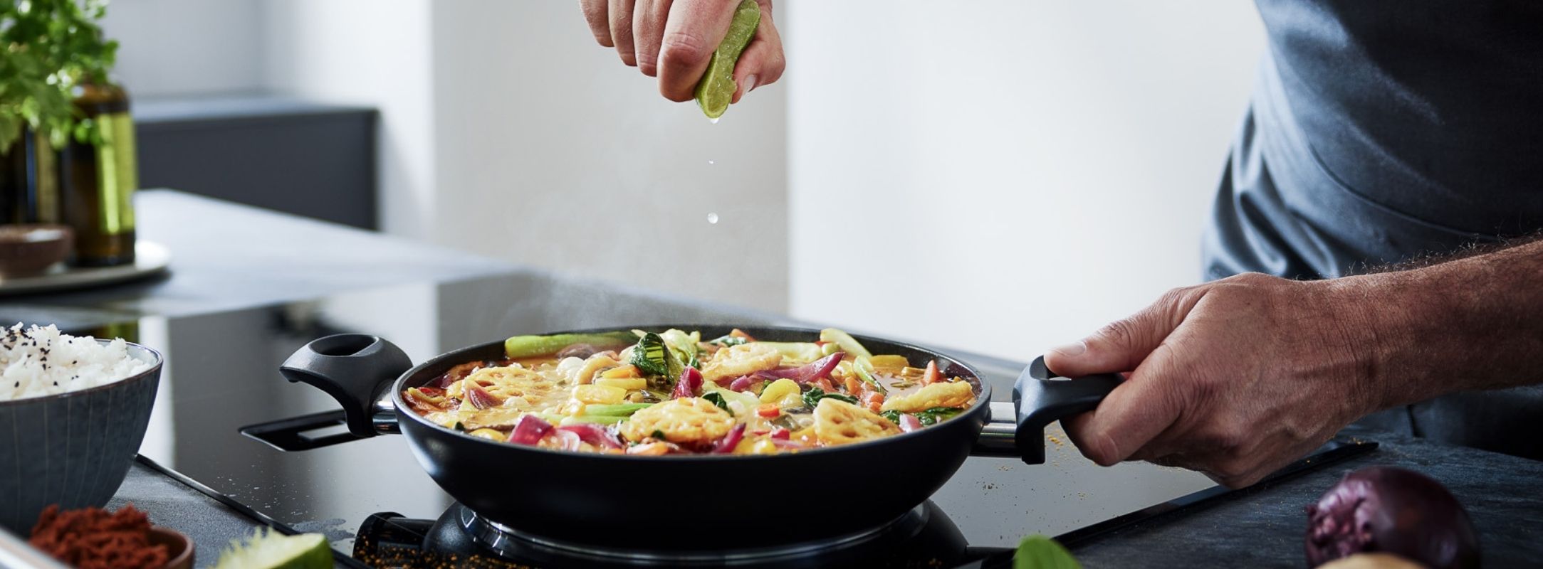 In einer Fissler-Pfanne auf dem Herd gibt man Limette zu lebhaftem Gemüse und Nudeln, dazu gibt es Reis und frische Produkte.