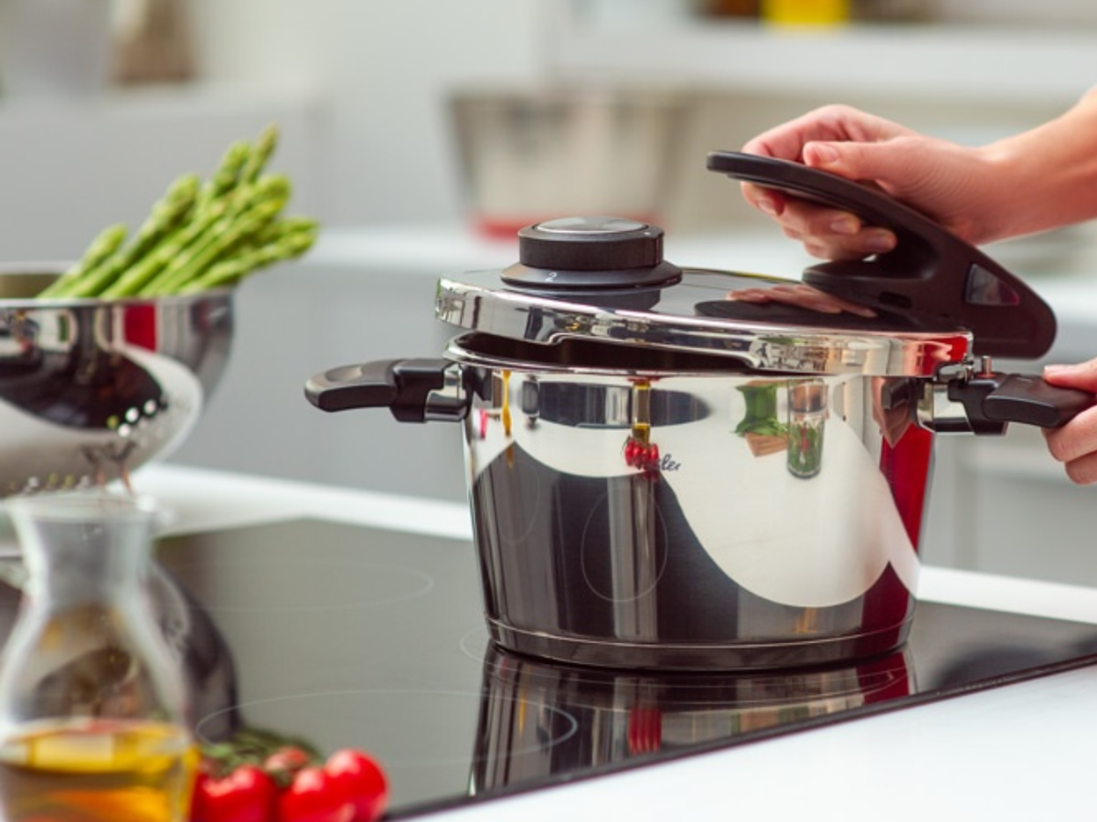 A person opens a Fissler stainless steel pressure cooker on a stovetop; asparagus, oil, and tomatoes complete the high-quality kitchen scene.