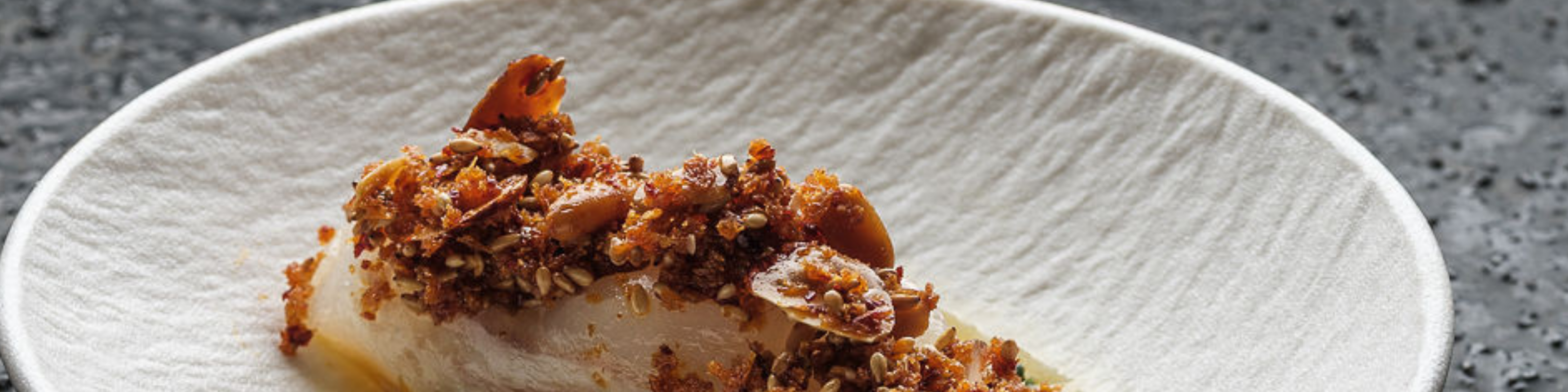 A white plate, elegantly placed on a textured gray surface, showcases a thoughtfully prepared dish topped with a crunchy blend of seeds and nuts. The sophisticated presentation reflects Fissler’s commitment to timeless design and precision engineering in kitchenware.