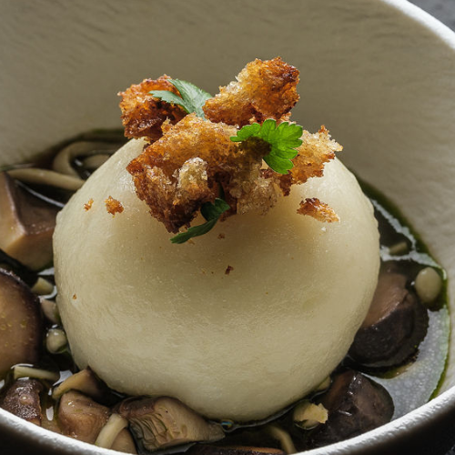 A perfectly round dumpling rests in savory mushroom broth, elegantly finished with golden crispy bread crumbs and a fresh sprig of parsley, served in a pristine white bowl—showcasing the refined presentation made possible with Fissler’s precision-engineered cookware.