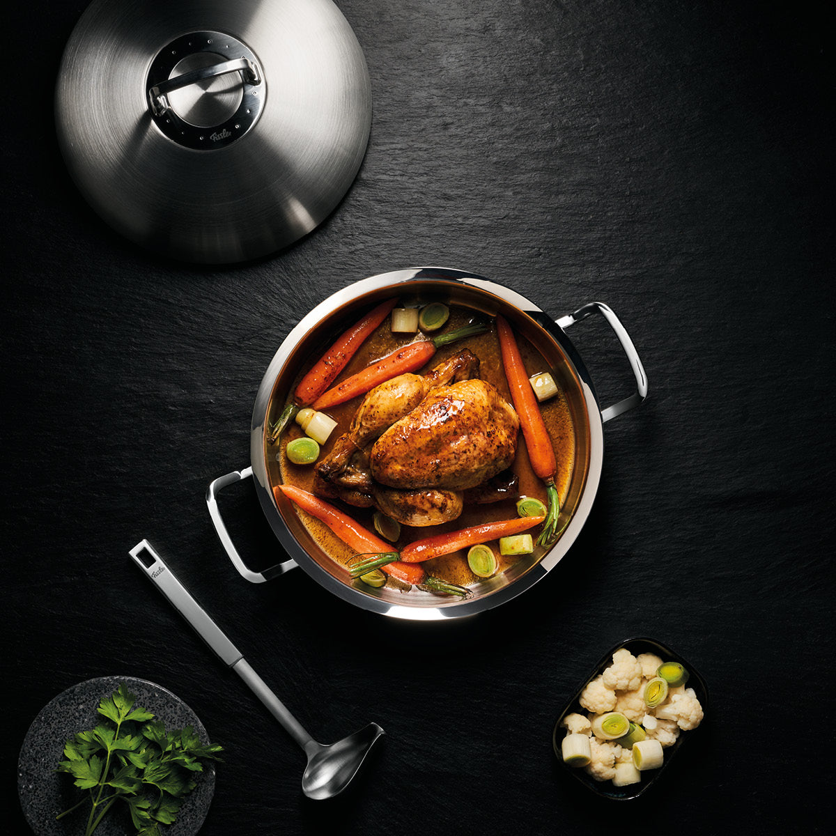 Ein gebratenes Hähnchen mit Gemüse in einem Fissler-Edelstahltopf, Deckel und Schöpflöffel daneben, Kräuter und Beilagen elegant arrangiert.