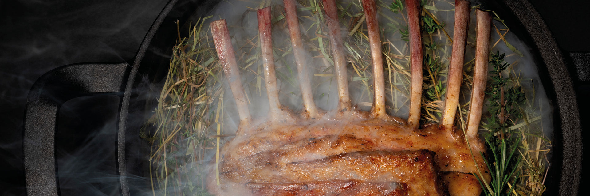 Ein gebratenes Lammkarree mit frischen Kräutern und Heu in einer gusseisernen Pfanne von Fissler, aromatischer Rauch unterstreicht das deutsche Kochgeschirr.