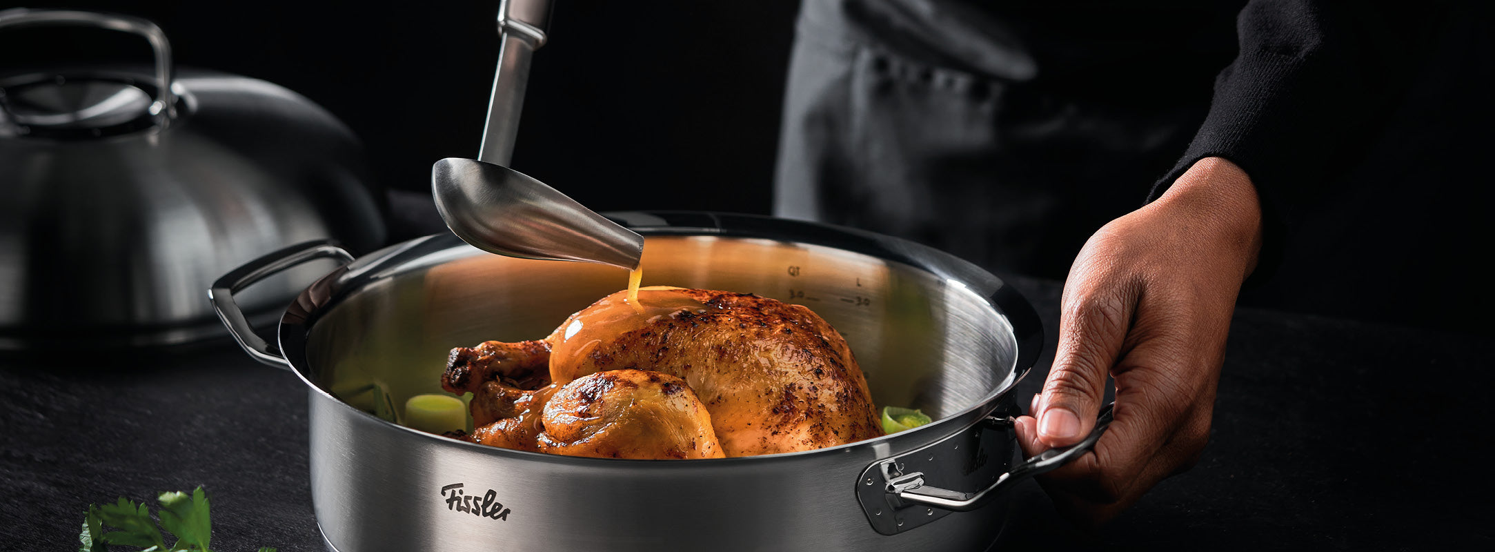 Die Soße wird über das Brathähnchen in einem Fissler-Edelstahltopf gegossen, mit Kräutern und Deckel in der Nähe - ein Beispiel für deutsche Qualitätskochgeschirre.