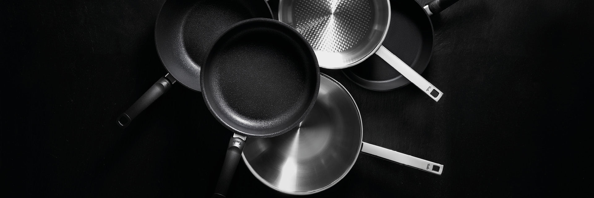 Eine Draufsicht auf Fissler Bratpfannen mit eleganten schwarzen und polierten Stahloberflächen, präzise gefertigt und kunstvoll auf einer dunklen Oberfläche angeordnet.