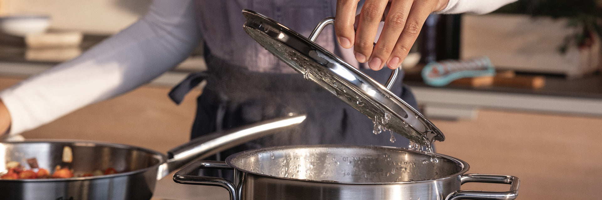 Eine Person hebt den Deckel eines Fissler-Edelstahltopfs auf dem Herd an, neben dem ein weiterer hochwertiger Topf in einer eleganten modernen Küche steht.