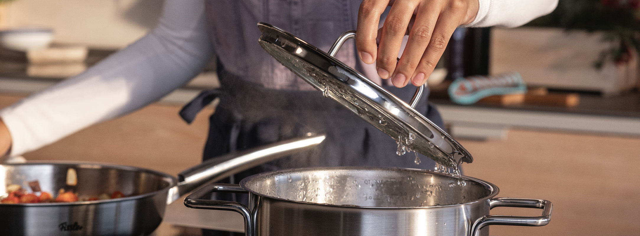Eine Person hebt den Deckel von einem dampfenden Fissler-Edelstahltopf auf dem Herd, neben dem eine Fissler-Pfanne in einer modernen Küche steht.