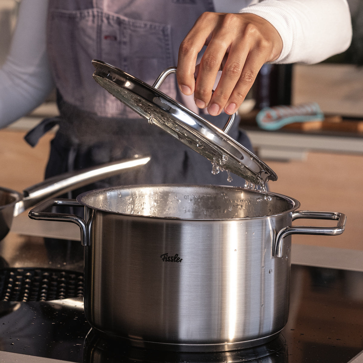 Eine Person hebt den Deckel von einem Fissler-Edelstahltopf auf dem Herd und gibt dampfendes, kochendes Wasser frei - Präzision Made in Germany".