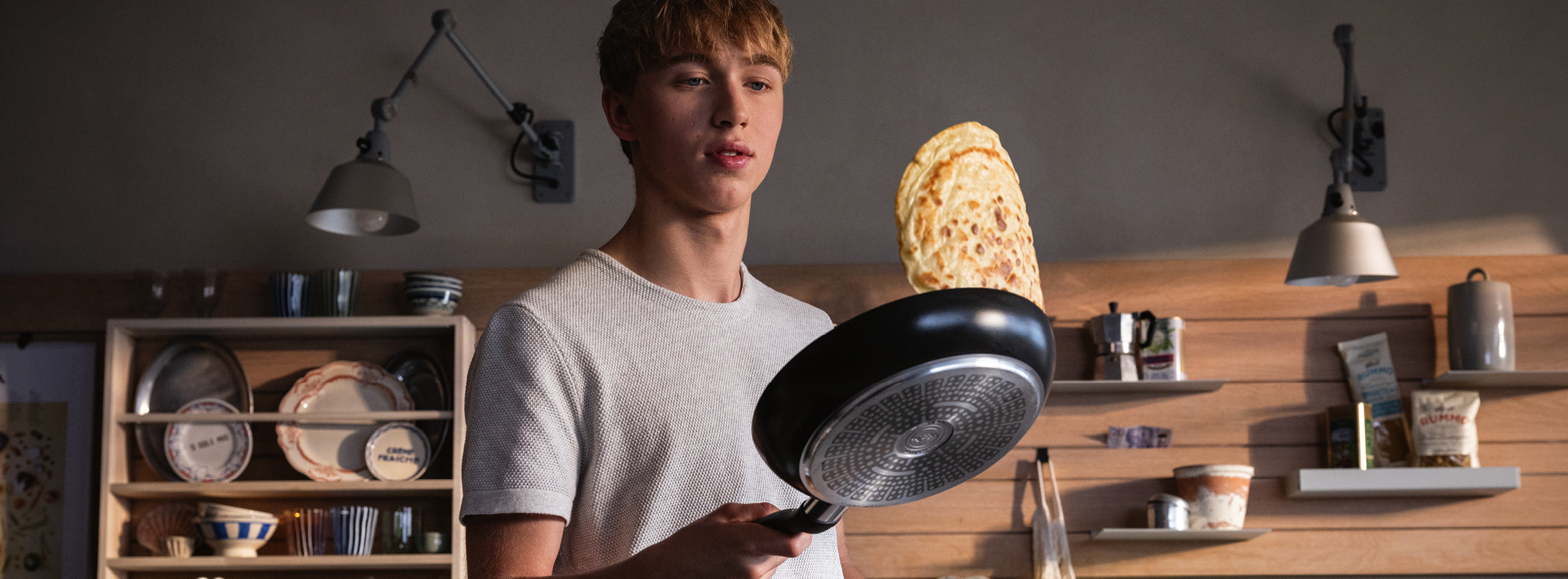 Ein junger Mann wendet einen Pfannkuchen in einer Fissler Bratpfanne, umgeben von hochwertigem Küchengeschirr auf Holzregalen in einer gemütlichen Küche.