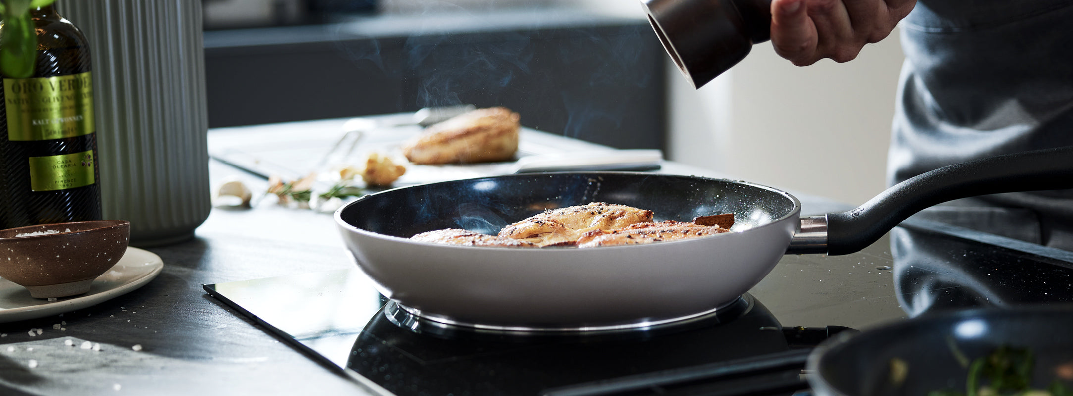 Ein Koch würzt Speisen in einer Fissler-Edelstahlpfanne auf dem Herd; Dampf steigt auf, während Öl und Kräuter in der Nähe sitzen, mit der Salzmühle in der Hand.