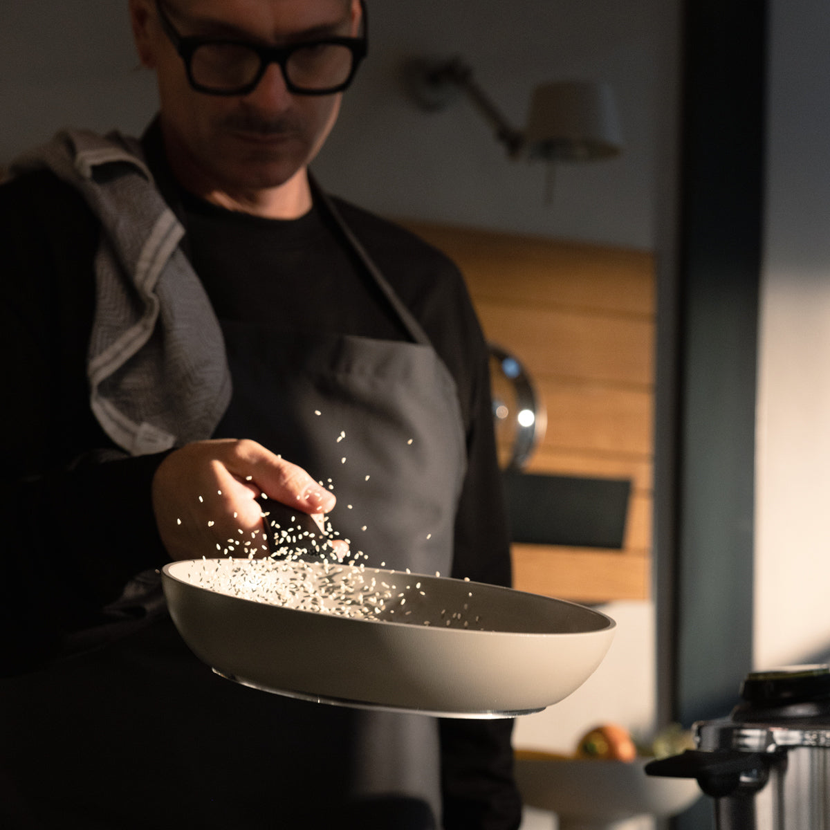 Eine Person mit Brille und Schürze schwenkt gekonnt Reis in einer Fissler-Pfanne, Körner in der Luft, die Küche erstrahlt in warmem Licht.