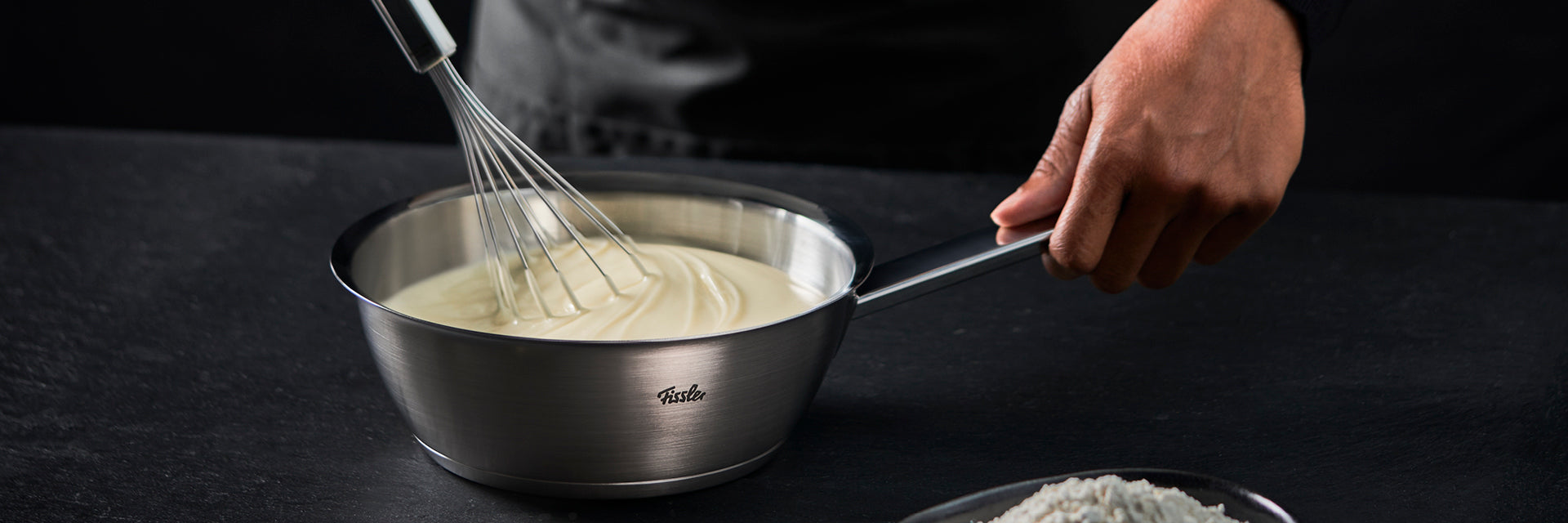 Eine Person rührt eine cremige Mischung in einem Fissler-Edelstahltopf auf einer dunklen Fläche; im Vordergrund steht eine Schüssel mit Mehl.