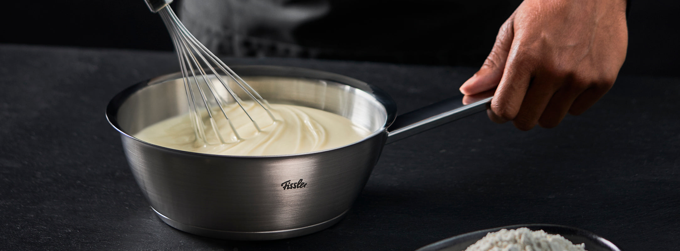 Eine Person verrührt weiße Soße in einem Fissler-Edelstahltopf auf einer dunklen Fläche, während Mehl in einer Schüssel daneben liegt.