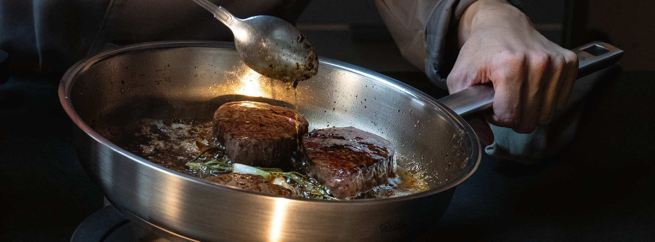 Eine Person brät Steaks mit Butter und Kräutern in einer Fissler-Edelstahlpfanne auf dem Herd und hält dabei den präzisionsgefertigten Griff fest.