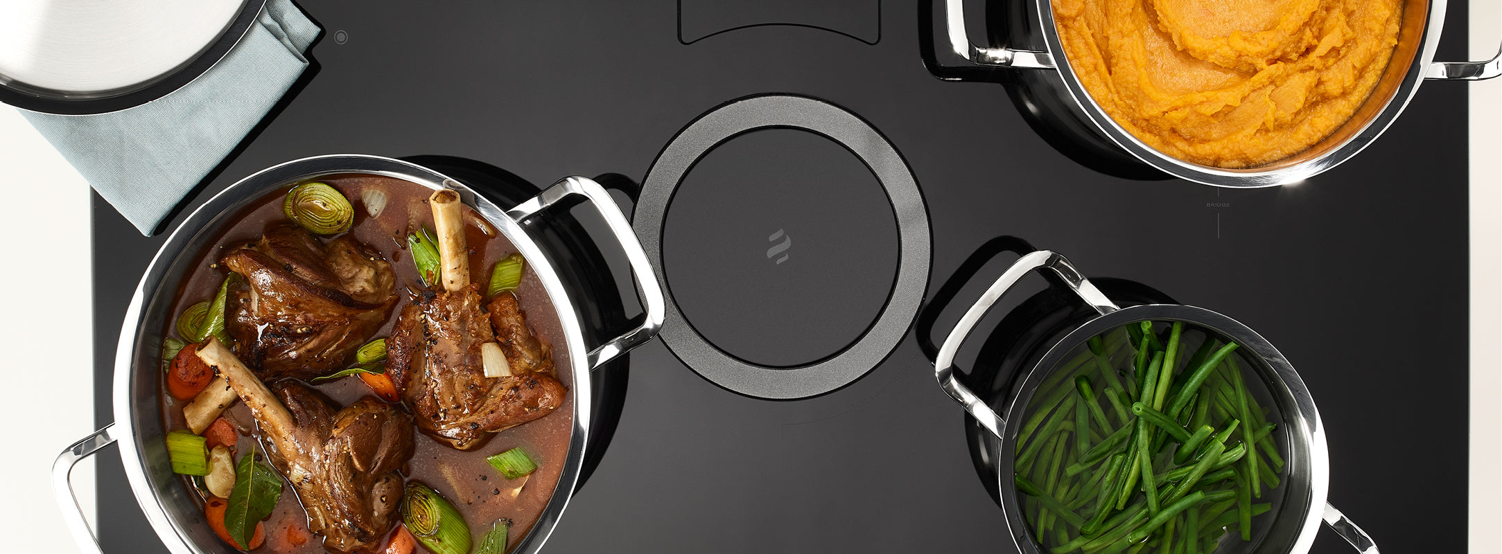 Blick von oben auf drei Fissler-Edelstahltöpfe auf einem schlichten schwarzen Herd, gefüllt mit Schmorbraten, grünen Bohnen und Süßkartoffeln.
