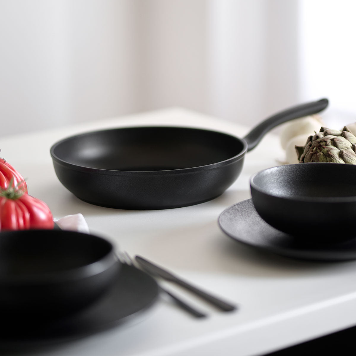 Fissler Cenit® Induktionspfanne mit Antihaftbeschichtung auf einem weißen Tisch, neben Küchenutensilien und frischem Gemüse in weichem Licht.