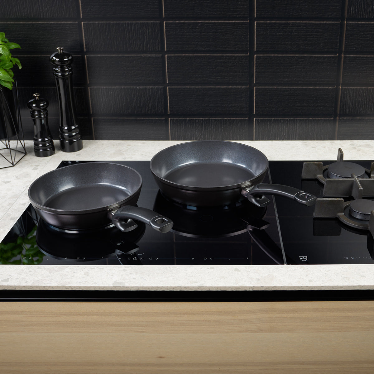 Two premium Fissler black frying pans, crafted with precision engineering and timeless German design, rest on a sleek glass stovetop. Nearby, elegant black pepper and salt grinders add sophistication, complemented by a touch of greenery and a modern black tile backsplash in the background.