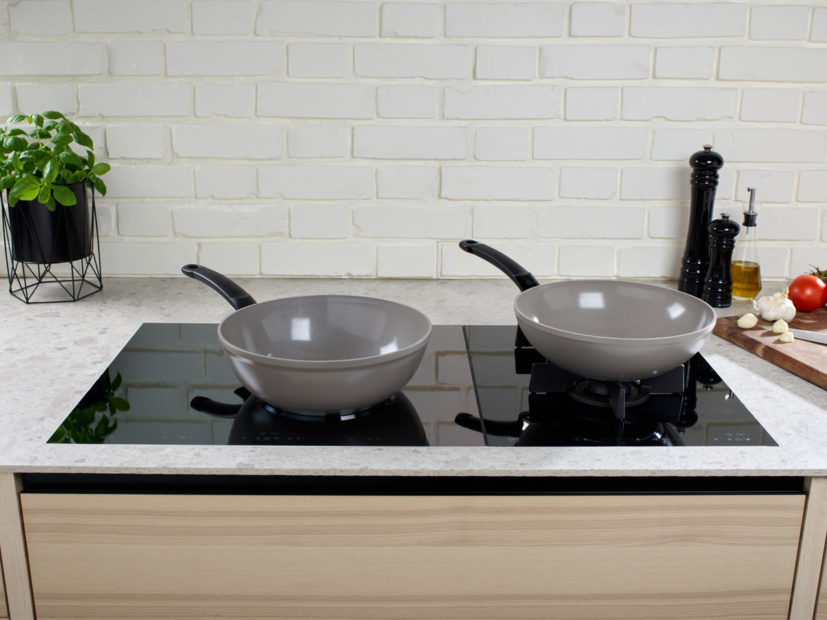 Fissler Ceratal® Wokpfanne und graue Bratpfanne auf schlichter schwarzer Herdplatte, umgeben von frischen Zutaten und einer weißen Backsteinplatte.