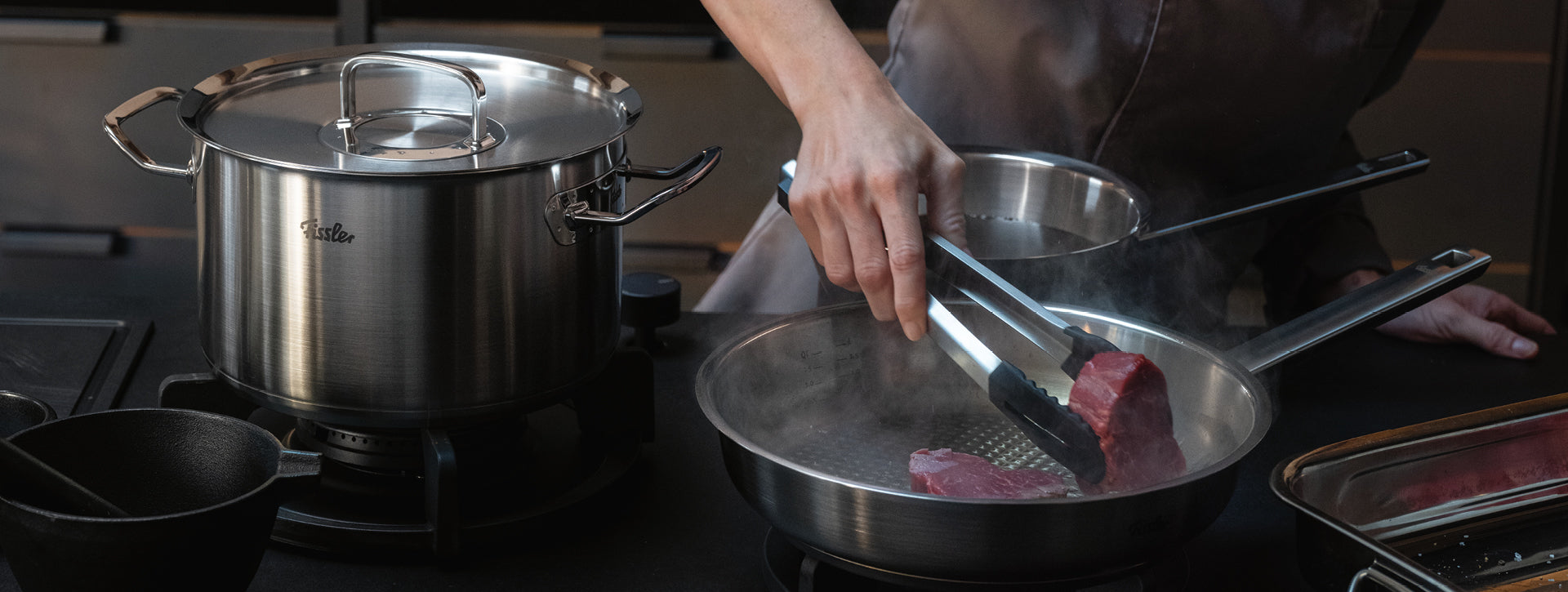 Ein Koch legt Steaks in eine Fissler-Edelstahlpfanne, die neben einem passenden Topf auf dem Herd steht. Präzision, Dampf und Innovation glänzen.