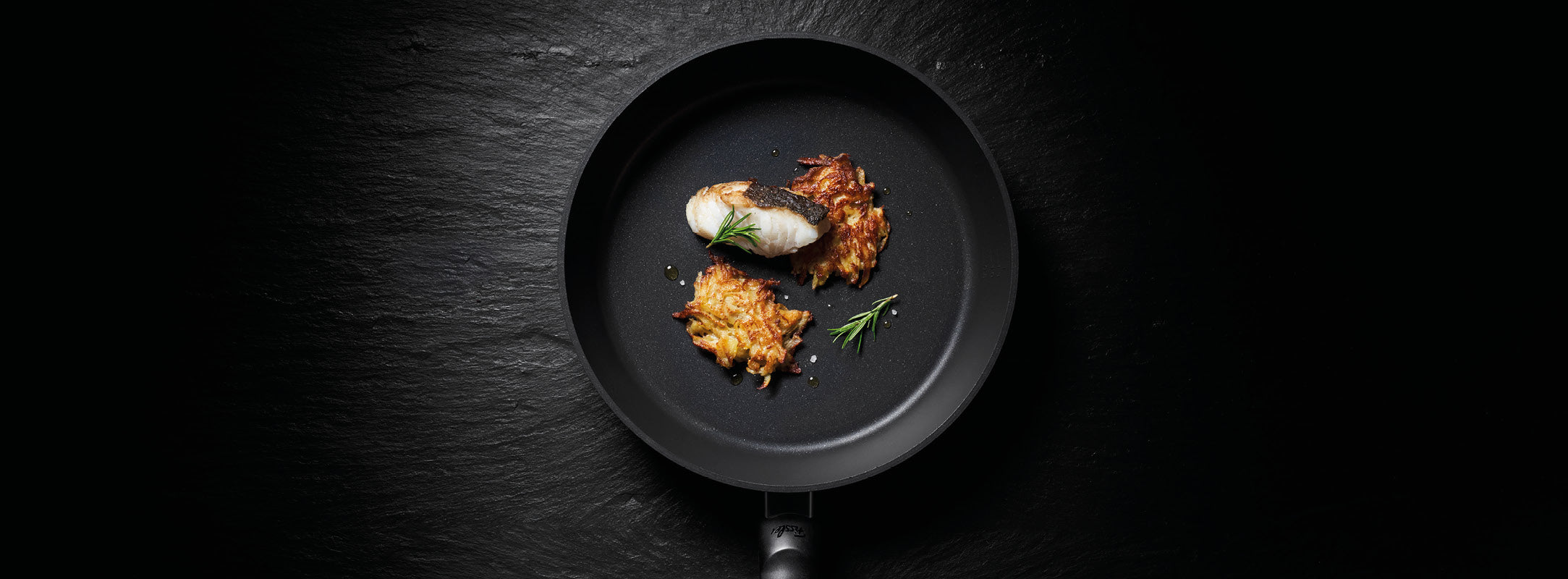Die schwarze Bratpfanne von Fissler präsentiert goldene Kartoffelpuffer und weißen Fisch mit Rosmarin, Eleganz und Präzision auf einer dunklen Oberfläche.